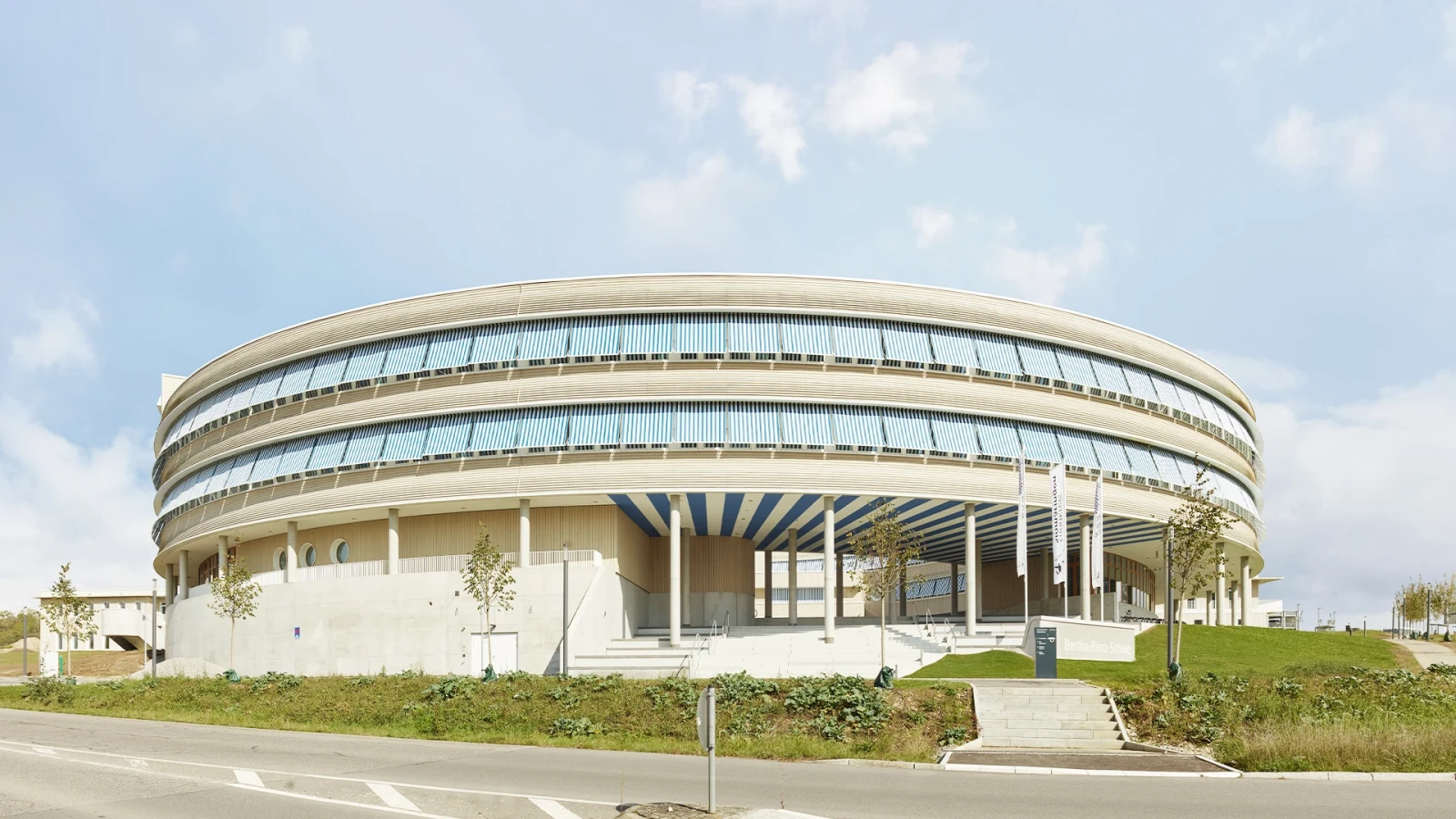 Die beiden Baukörper prägen naturfarbene Holzfassaden, weiße Akzente und gezielt gesetzte Öffnungen. Geschützte Holzfenster und blau-weiße Markisen verleihen dem Ensemble Leichtigkeit und Charakter.